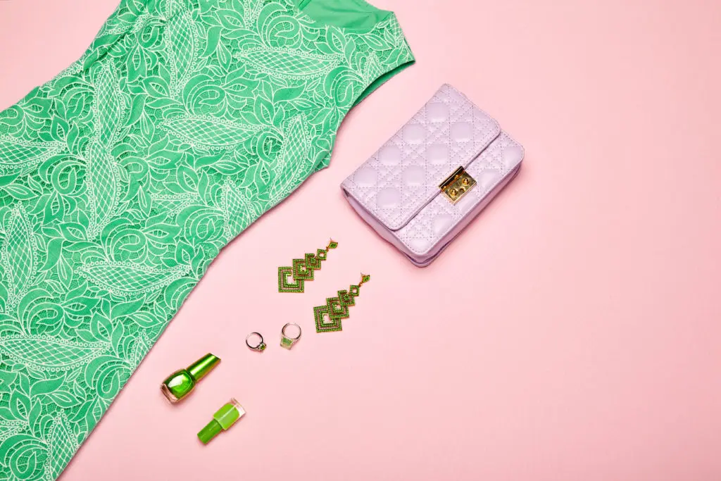 flat lay of green gemstone earrings and rings with a pink purse and green dress on a pink background