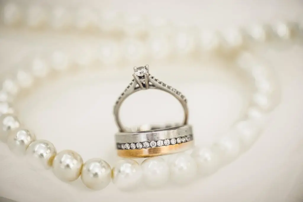 diamond engagement ring set next to pearl necklace on a white background