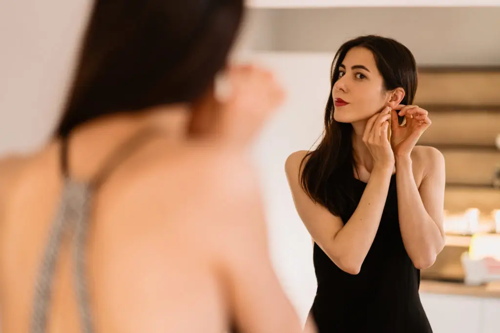 woman trying on jewelry for a fancy evening in front of a mirror wearing a black dress