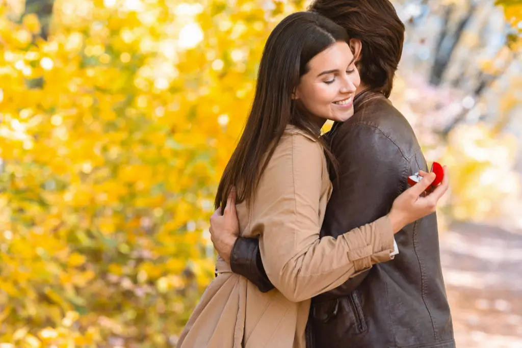 woman embraces a man while holding an engagement ring with a yellow leaves behind them celebrating a fall proposal