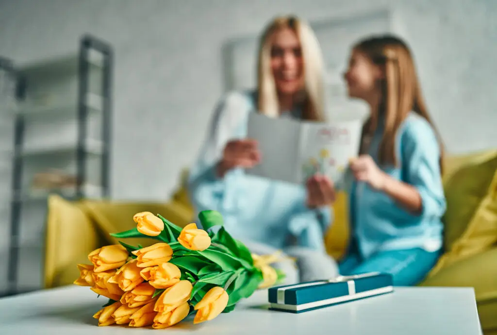 yellow tulips and a jewelry box sit on a table with a mother and daughter opening a Mother's Day card in the background