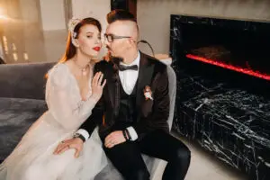 An edgy non traditional bride and groom pose sitting on a couch in front of a fireplace